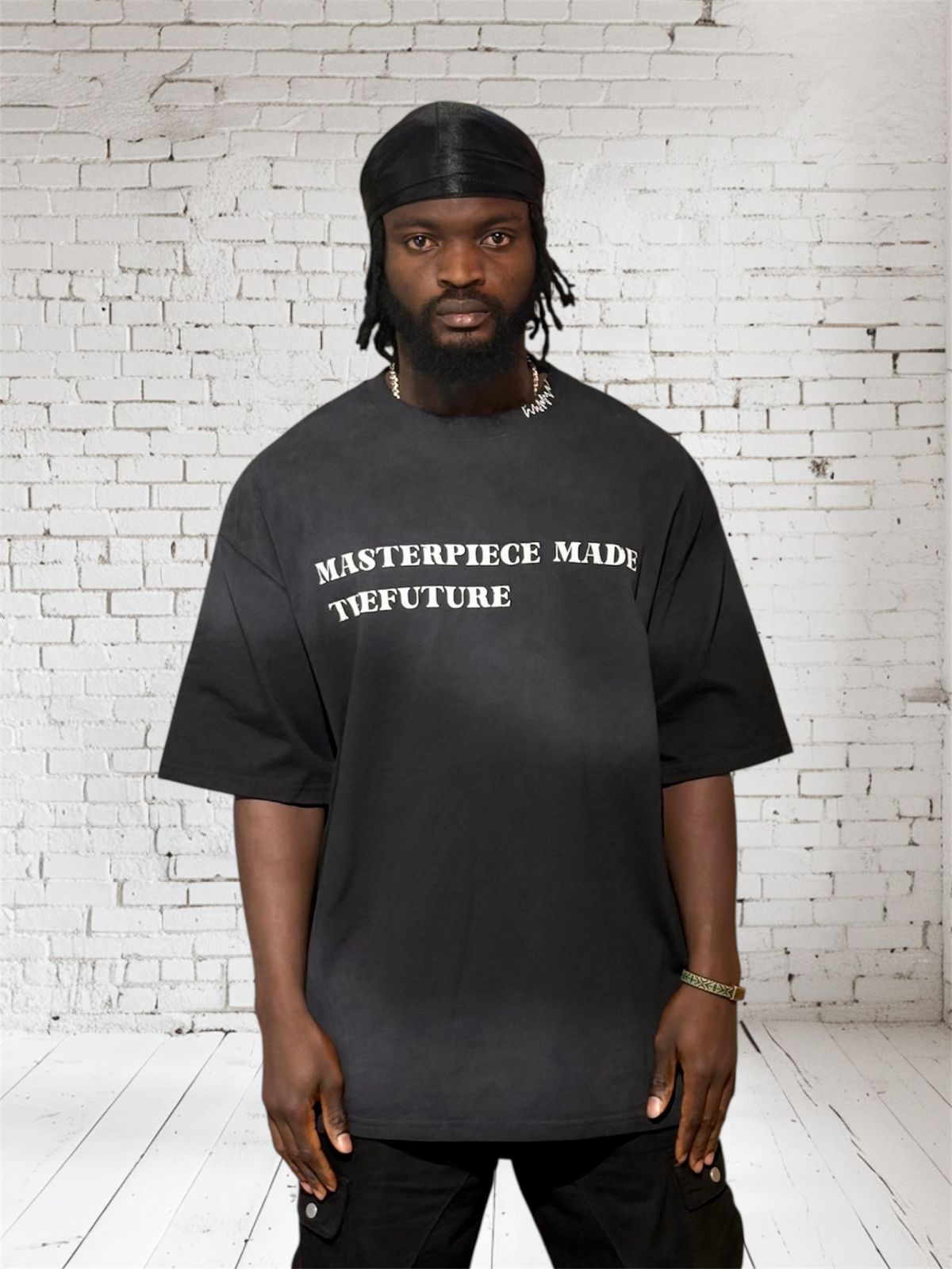 Person wearing a black t-shirt with 'MASTERPIECE MADE TTFUTURE' text against a white brick wall.